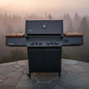 A black gas grill with wooden side shelves, upgraded with BBQ Boards® Napoleon Prestige 500 Connected Side Cutting Boards (Matching Pair), stands on a stone patio, framed by a misty forest and tall trees in the background.