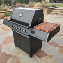 A black outdoor gas grill with four control knobs and two wooden side shelves—one upgraded with BBQ Boards® Napoleon Prestige 500 Connected Side Cutting Boards (Matching Pair) by BBQ Boards®—sits on caster wheels on a stone patio in a landscaped backyard.