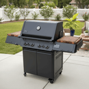 A Napoleon Prestige 500 grill features BBQ Boards® Connected Side Cutting Boards (Matching Pair) on its wooden side shelves, positioned on a patio with greenery and white fencing in the backyard.