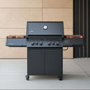 A black outdoor gas grill with four knobs, closed lid, wheels, and BBQ Boards® Napoleon Prestige 500 Connected Side Cutting Boards by BBQ Boards® on the side shelves stands on a patio against a beige tiled wall.