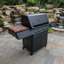 A large black gas grill with wooden side shelves stands on a stone patio, upgraded with BBQ Boards® Napoleon Prestige 500 Connected Side Cutting Boards (Matching Pair) by BBQ Boards®, set against rocks, plants, and a waterfall.