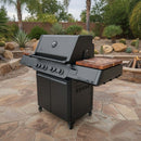 A large black outdoor grill with multiple knobs, a built-in thermometer, and a matching pair of BBQ Boards® Napoleon Prestige 500 Connected Side Cutting Boards sits on a stone patio amid desert landscaping.