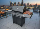 A large black gas grill with four knobs stands on a tiled balcony at dusk, overlooking a city skyline. A BBQ Boards® Napoleon Prestige 500 Connected Side Cutting Board by BBQ Boards® rests on the BBQ side shelf, ready for evening grilling.
