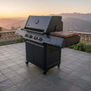A BBQ Boards® Napoleon Prestige 500 Connected Side Cutting Board by BBQ Boards® is on a tiled patio, featuring modern black grilling style with wooden side shelves, sunset mountain views, stone railing, and greenery in the background.