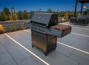 A large black outdoor gas grill with multiple burners features a BBQ Boards® Napoleon Prestige 500 Connected Side Cutting Board, set on a modern patio with built-in lighting and stylish outdoor seating in the background.