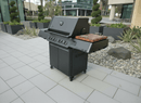 A sleek black gas grill with a built-in thermometer stands on a tiled patio. On its side shelf sits the BBQ Boards® Napoleon Prestige 500 Connected Side Cutting Board by BBQ Boards®. Planters, rocks, and office buildings are in the background.