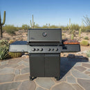 A BBQ Boards® Napoleon Prestige 500 Connected Side Cutting Board is attached to a black gas grill with four knobs and side shelves, set on a stone patio amid desert cacti under a clear blue sky.