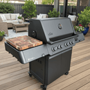 A large black outdoor gas grill with four knobs and a thermometer sits on a wooden deck, next to a BBQ Boards® Napoleon Prestige 500 Connected Side Cutting Board. Patio furniture and potted plants are in the background.
