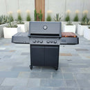 A modern black gas grill with four knobs and side shelves stands on a stone patio, featuring the BBQ Boards® Napoleon Prestige 500 Connected Side Cutting Board, surrounded by white planters with tall green plants by a concrete wall.