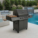 A BBQ Boards® Napoleon Prestige 500 Connected Side Cutting Board by BBQ Boards® is attached to a sleek black outdoor gas grill on a patio next to a pool, surrounded by lounge furniture and greenery.
