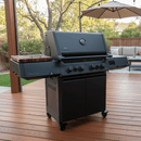 A large black gas grill with four knobs, side burner, and storage cabinet stands on a deck outdoors. On its side shelf is a BBQ Boards® Napoleon Prestige 500 Connected Side Cutting Board. Trees, fence, and patio furniture are visible in the background.
