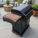 A black outdoor gas grill with four knobs and a BBQ Boards® Napoleon Rogue PRO Griddle Side Cutting Board sits on a patio, surrounded by potted plants, a wooden coffee table, and outdoor seating.