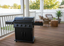 A black outdoor gas grill with a BBQ Boards® Napoleon Rogue PRO Griddle Side Cutting Board sits on a wooden deck with wicker chairs, a round table, and a potted plant, surrounded by white railings and greenery.