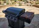 A black outdoor gas grill with four knobs and a BBQ Boards® Napoleon Rogue PRO Griddle Side Cutting Board by BBQ Boards® stands on a stone patio, surrounded by rocks, desert plants, and cacti in a landscaped backyard.