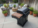 A sleek black grill with a BBQ Boards® Napoleon Rogue PRO Griddle Side Cutting Board stands on the patio near outdoor seating, potted plants, a coffee table, and a pool, all enclosed by a wooden fence.