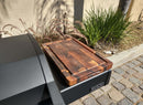 The BBQ Boards® Napoleon Rogue PRO Griddle Side Cutting Board by BBQ Boards® sits on a black outdoor grill’s side shelf next to a cobblestone path, with lush green plants lining a sunlit beige wall.