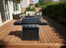 A BBQ Boards® Napoleon Rogue PRO Griddle Side Cutting Board by BBQ Boards® is on a black outdoor gas grill with four knobs on a wooden deck. Patio furniture sits nearby, in front of a beige house amid green shrubs and trees.