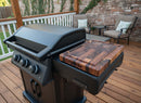 A BBQ Boards® Napoleon Rogue PRO Griddle Side Cutting Board rests on a modern gas grill with three knobs on a wooden deck. Two cushioned patio chairs and an ivy-clad brick wall in the background create an inviting gathering space.