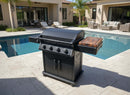 A BBQ Boards® Napoleon Rogue PRO Griddle Side Cutting Board by BBQ Boards® is on a black four-burner outdoor gas grill with a wooden side tray on a tile patio by the pool, surrounded by patio furniture and a modern house in the background.