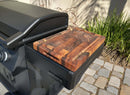 A close-up of a BBQ Boards® Napoleon Rogue PRO Griddle Side Cutting Board resting on a black grill's side shelf, with stone pavers and lush green plants in the background—perfect for any BBQ Boards® fan.