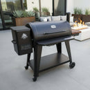 A BBQ Boards® Pit Boss 1150DX Side Cutting Board by BBQ Boards® is mounted on a modern black pellet grill and smoker with a digital control panel, positioned on a patio near outdoor seating, a fire pit, and potted plants.