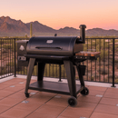 A BBQ Boards® Pit Boss 1150DX Side Cutting Board is shown attached to a black pellet grill on a tiled patio, with desert plants and mountains at sunset, all framed by a black metal fence.