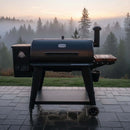 A black Pit Boss 1150DX grill with a BBQ Boards® Pit Boss 1150DX Side Cutting Board stands on wet patio tiles, overlooking misty pine trees and a sunrise sky.