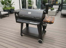A black Pit Boss 1150DX pellet grill with a smokestack and wood shelf is on a modern deck, alongside outdoor furniture, potted plants, and the BBQ Boards® Pit Boss 1150DX Side Cutting Board by BBQ Boards® for easy meal prep.
