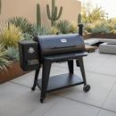 A black Pit Boss 1150DX grill stands on a modern patio with desert plants. A BBQ Boards® Pit Boss 1150DX Side Cutting Board by BBQ Boards® rests on the side board, ready for your next cookout near built-in seating and a beige wall.