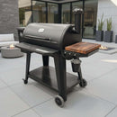 A BBQ Boards® Pit Boss 1150DX Side Cutting Board is attached to a black Pit Boss 1150DX pellet grill with utensil hooks and chimney, placed on gray patio tiles amid modern outdoor furniture and fire pits.