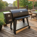 A BBQ Boards® Pit Boss 1150DX Side Cutting Board is on a black pellet grill and smoker on a wooden deck, surrounded by patio furniture and greenery as sunlight streams through the trees.