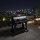 A BBQ Boards® Pit Boss 1150DX Side Cutting Board is attached to a black pellet grill on a rooftop patio at night, with a fire pit, modern seating, lush plants, and an illuminated city skyline in the background.