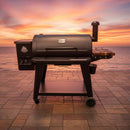 A BBQ Boards® Pit Boss 1150DX Side Cutting Board by BBQ Boards® is displayed on the Pit Boss 1150DX pellet grill's side board on a tiled patio, ocean views and a glowing sunset sky in the background.