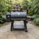 A BBQ Boards® Pit Boss 1150DX Side Cutting Board is set on a black Pit Boss 1150DX pellet smoker grill on a stone patio amid tropical plants, with a multi-tiered waterfall and fountain in the background, ready for grilling.