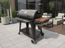 A BBQ Boards® Pit Boss 1150DX Side Cutting Board is attached to a black pellet grill with wheels, shown on a patio featuring potted plants and outdoor seating in the background.