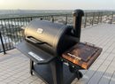 A black outdoor smoker grill with a BBQ Boards® Pit Boss 850DX Side Cutting Board sits on a rooftop deck with metal railings, overlooking the city skyline and river.