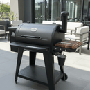A black outdoor pellet grill with a chimney and a BBQ Boards® Pit Boss 850DX Side Cutting Board is on a modern patio with gray tiles and white cushioned patio furniture in the background.