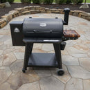 A black outdoor pellet grill and smoker with a BBQ Boards® Pit Boss 850DX Side Cutting Board stands on a stone patio, featuring a side hopper, chimney, and small wooden shelf amid stone walls and lush greenery in the background.