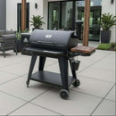 A black outdoor pellet grill with a BBQ Boards® Pit Boss 850DX Side Cutting Board and wood shelf stands on light tile patio near a modern house, surrounded by potted plants and patio furniture.