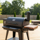 A BBQ Boards® Pit Boss 850DX Side Cutting Board is attached to a black pellet grill on a wooden deck, with a fenced yard and green trees in the background under bright sunlight.
