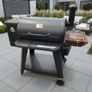 A black Pit Boss pellet grill with a smoking chamber, chimney, and a BBQ Boards® Pit Boss 850DX Side Cutting Board by BBQ Boards®, set on a modern patio with stylish furniture and plants in the background.
