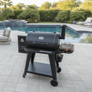 A BBQ Boards® Pit Boss 850DX Side Cutting Board is on a black pellet grill, set on a stone pool deck beside a blue swimming pool, surrounded by lounge chairs and greenery under the sun.