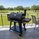 A black outdoor grill with a side smoker box and a BBQ Boards® Pit Boss 850DX Side Cutting Board stands on a tiled patio overlooking a golf course, green grass, and trees under a clear blue sky.