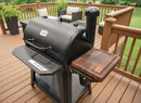 A black outdoor pellet grill with a BBQ Boards® Pit Boss 850DX Side Cutting Board and a wooden side shelf sits on a brown wooden deck, surrounded by patio furniture and green plants in the background.