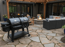 A BBQ Boards® Pit Boss Pellet Gas Combo Side Cutting Board sits on a stone patio with a fire pit, Adirondack chairs, and wicker sofa set with blue cushions, surrounded by string lights and potted plants.