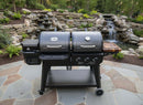 A BBQ Boards® Pit Boss Pellet Gas Combo Side Cutting Board sits on a black combo grill with multiple compartments and controls, placed on a stone patio before a landscaped garden with a rock waterfall and fountain in the background.