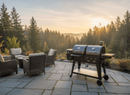 A patio with wicker chairs and a fire pit overlooks a forested valley at sunrise. A BBQ Boards® Pit Boss Pellet Gas Combo Side Cutting Board by BBQ Boards® rests on the stone patio, framed by tall pines and misty sunlight.