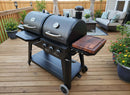 A BBQ Boards® Pit Boss Pellet Gas Combo Side Cutting Board by BBQ Boards® is attached to a dual-chamber black grill on a wooden deck, surrounded by potted plants and patio furniture.