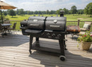 A BBQ Boards® Pit Boss Pellet Gas Combo Side Cutting Board is attached to a large black grill with two compartments and control knobs on a wooden deck overlooking a golf course, with patio furniture, trees, and blue sky in the background.