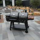A BBQ Boards® Pit Boss Pellet Gas Combo Side Cutting Board is featured on a black dual-chamber grill with thermometer and side burner, standing on a stone-tiled patio with chairs, rocks, plants, and a building in the background.